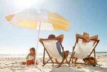 parents sit in beach chairs while child plays in sand under umbrella on sunny day
