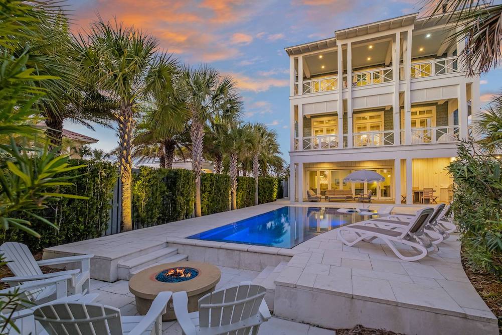 backyard of a beach rental featuring lines of palm trees, a recessed fire pit and infinity pool