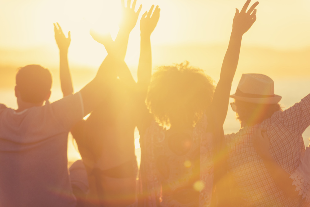 group of friends at beach during sunset, backs turned, and arms raised in celebration 